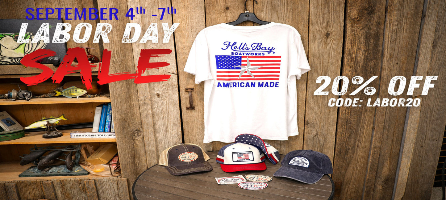 A white graphic T-shirt hanging on a black hanger against a rustic wooden wall, with sale text overlays reading 'September 4th -7th Labor Day Sale' and '20% Off Code: Labor20'. Several baseball caps and patches are displayed on a wooden table below.