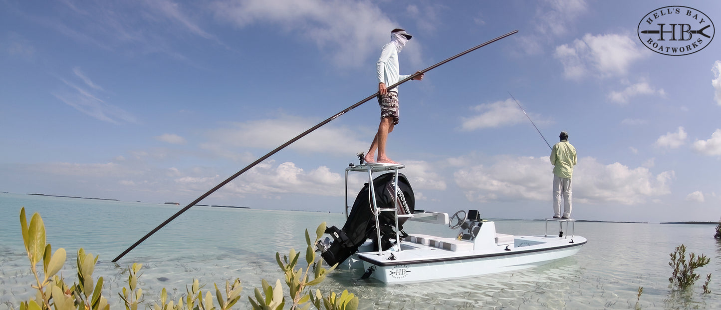 A white fishing boat on calm turquoise water with two people on board, one standing and holding a long pole and one standing further back holding a fishing rod. Low green plants frame the bottom of the scene under a blue sky with scattered clouds.