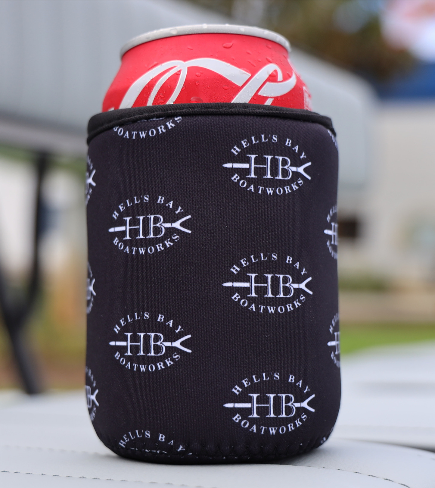 Black Hell’s Bay neoprene can cooler with repeating white HB Boatworks logos, holding a red soda can.